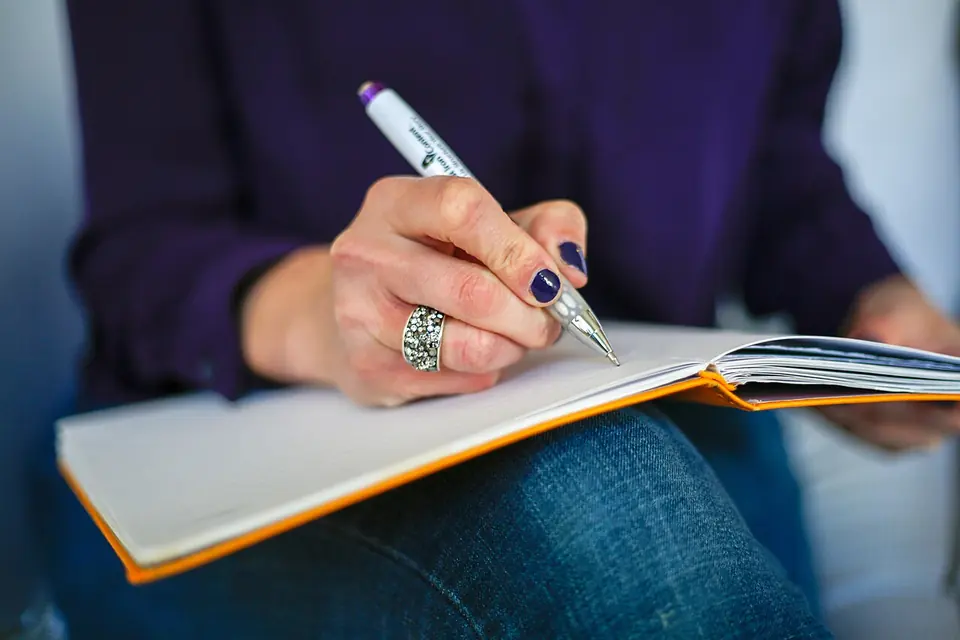 Main qui écrit au stylo dans un carnet ouvert posé sur ses genoux